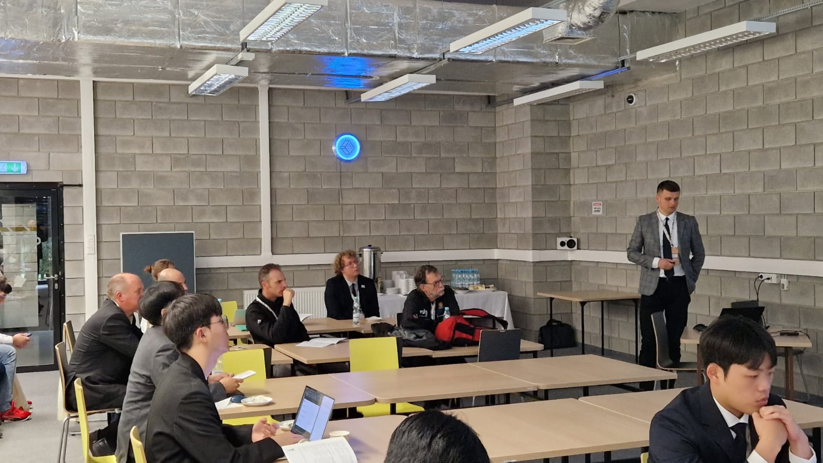 Fot. 1. Dariusz Kalinko podczas wygłaszania prezentacji multimedialnej stojąc na środku sali przodem do ekranu multimedialnego, obok zgromadzonych słuchaczy.