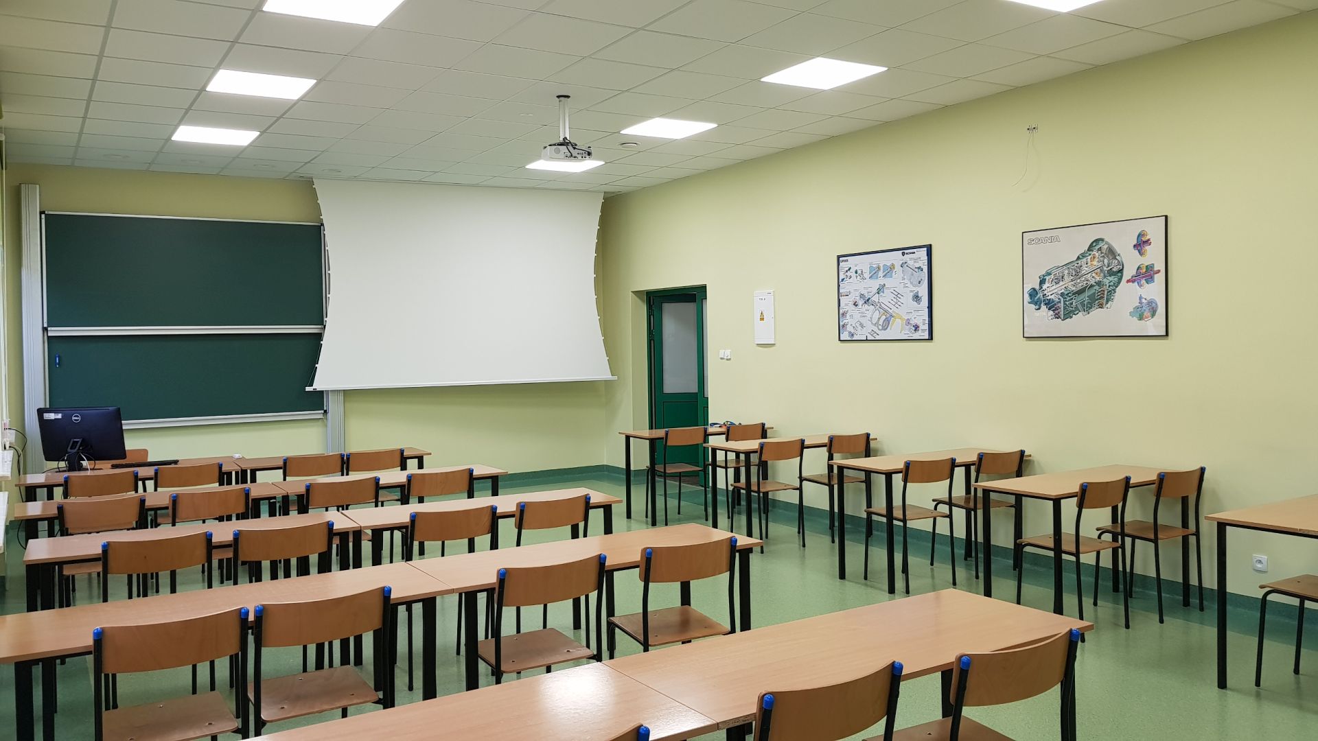 Fot. 1. Ogólny widok sali wykładowej nr 2 po remoncie, na pierwszym planie ławki i krzesła, na dalszym planie tablica akademicka, projektor multimedialny i ekran projekcyjny, na ścianach po prawej plakaty dydaktyczne.