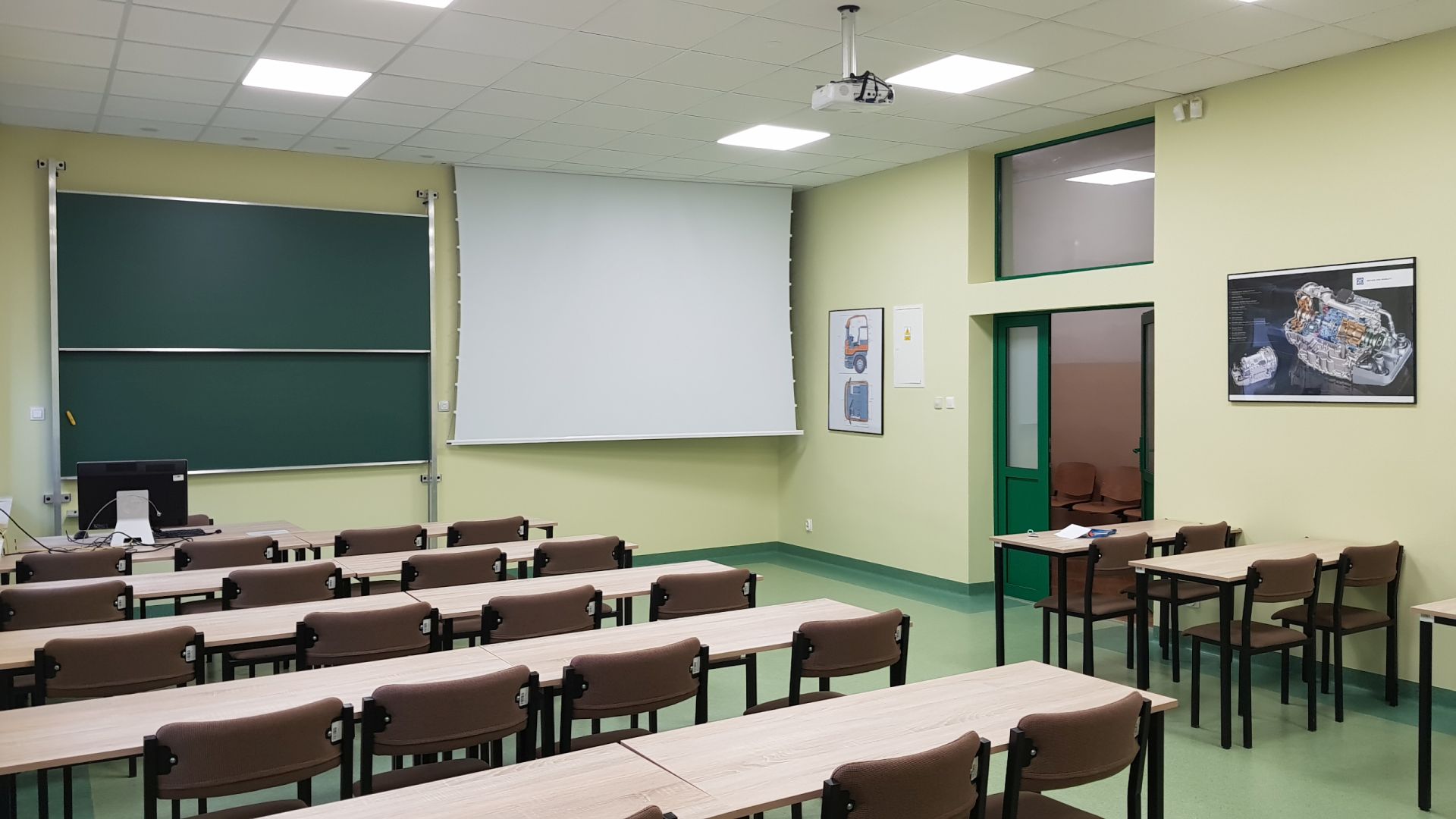 Fot. 2. Ogólny widok sali wykładowej nr 5 po remoncie, na pierwszym planie ławki i krzesła, na dalszym planie tablica akademicka, projektor multimedialny i ekran projekcyjny, na ścianach po prawej plakaty dydaktyczne.