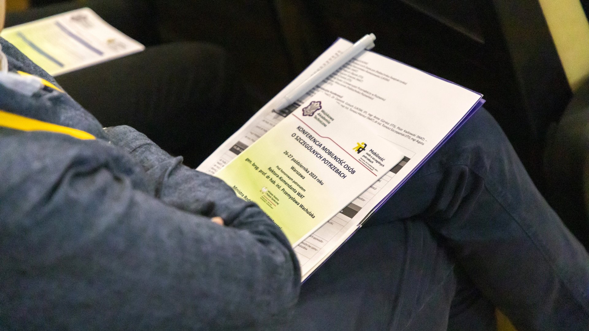 Fot. 6. Uczestnik siedzący na sali konferencyjnej z ulotką papierową na kolanach.