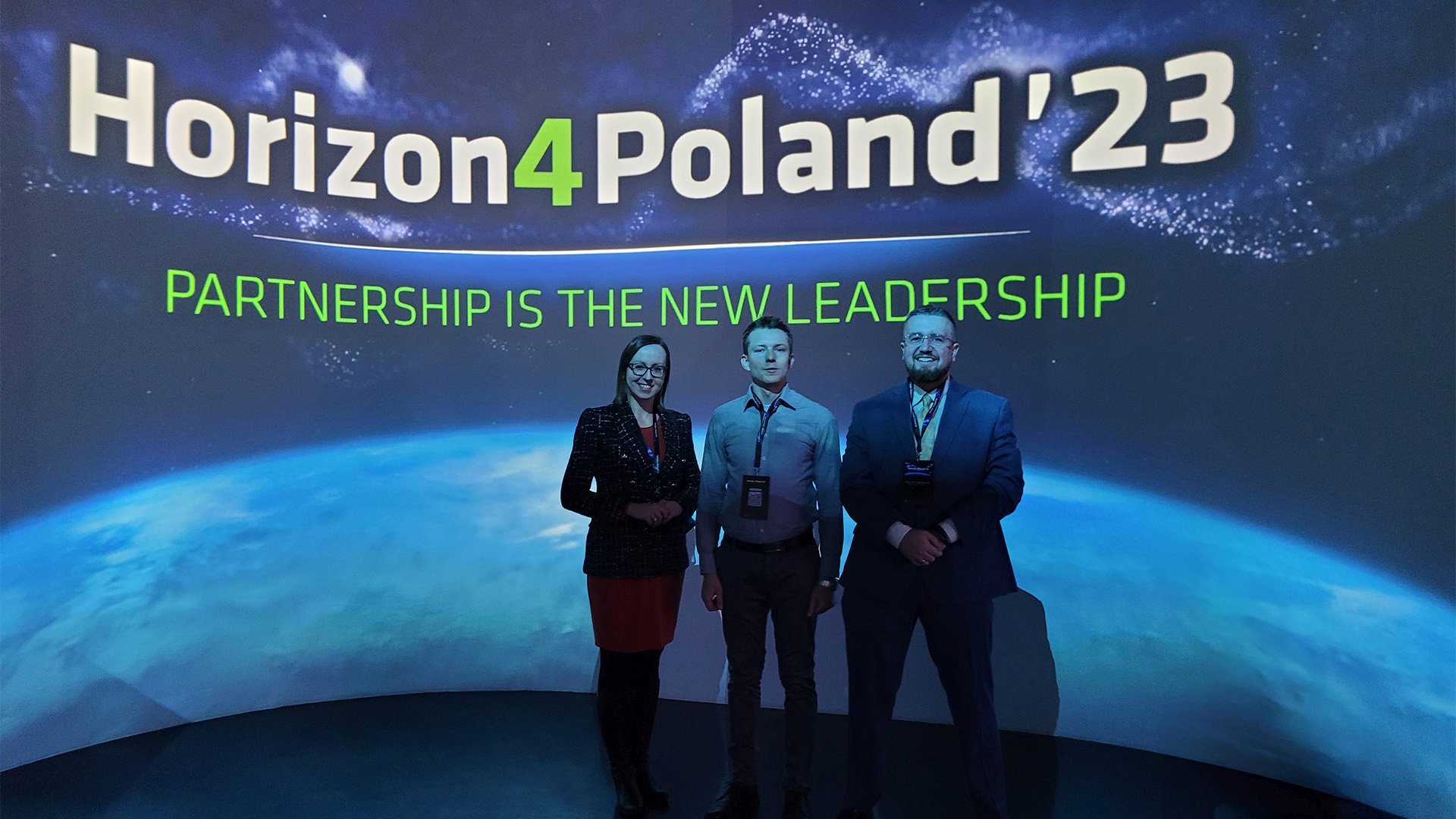 Fot. 1. Kpt. dr inż. Adrian Trzeciak z 2 innymi osobami stojacy na tle niebieskiego baneru z napisem Horizon4Poland'2023'