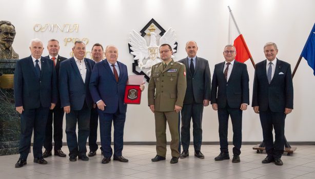 Grupa osób stojących obok siebie, przedstawiciele WAT i Pronar. W tle godło WAT i flaga Polski.