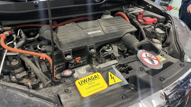 Widok komory silnikowej samochodu Honda CRV po podniesieniu pokrywy – na pierwszym planie widoczna tabliczka ostrzegawcza informująca o wykonywania prac elektrycznych przy instalacji wysokiego napięcia