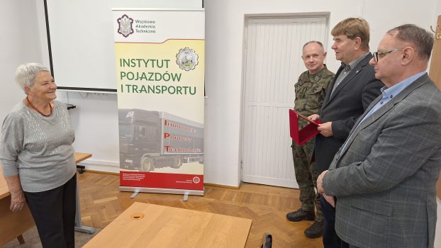 Doktor habilitowany inżynier Mirosław Gidlewski, dyrektor Instytutu Pojazdów i Transportu stojący w gronie zastępców, wygłaszający okolicznościowe przemówienie – po lewej stronie pani Wiesława Zielnik, na dalszym planie rollup z nazwą i logo instytutu