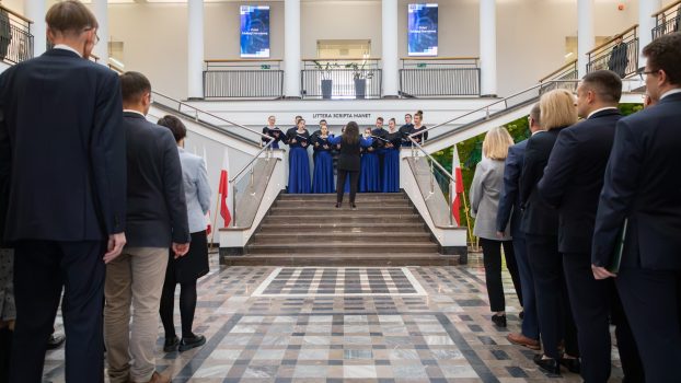 Na schodach stoi chór akademicki. Osoby w takich samych czarno-niebieskich strojach. Na pierwszym planie tyłem odznaczone osoby.
