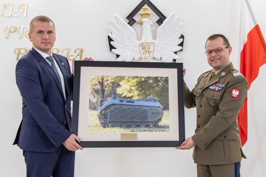 Rektor-Komendant WAT oraz Prezes firmy Milrem Robotics pozują do zdjęcia wspólnie trzymając w rękach okolicznościowy upominek (oprawione zdjęcie flagowego produktu firmy - bezzałogowy pojazd lądowy THeMIS) od Firmy Milrem Robotics dla WAT.