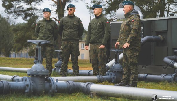 Podchorążowie przy początkowym punkcie rurociągu. Grupa żołnierzy stoi na dworzu przy rurociągi.