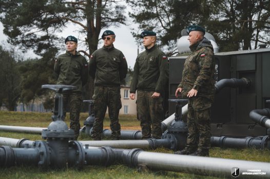 Podchorążowie przy początkowym punkcie rurociągu. Grupa żołnierzy stoi na dworzu przy rurociągi.