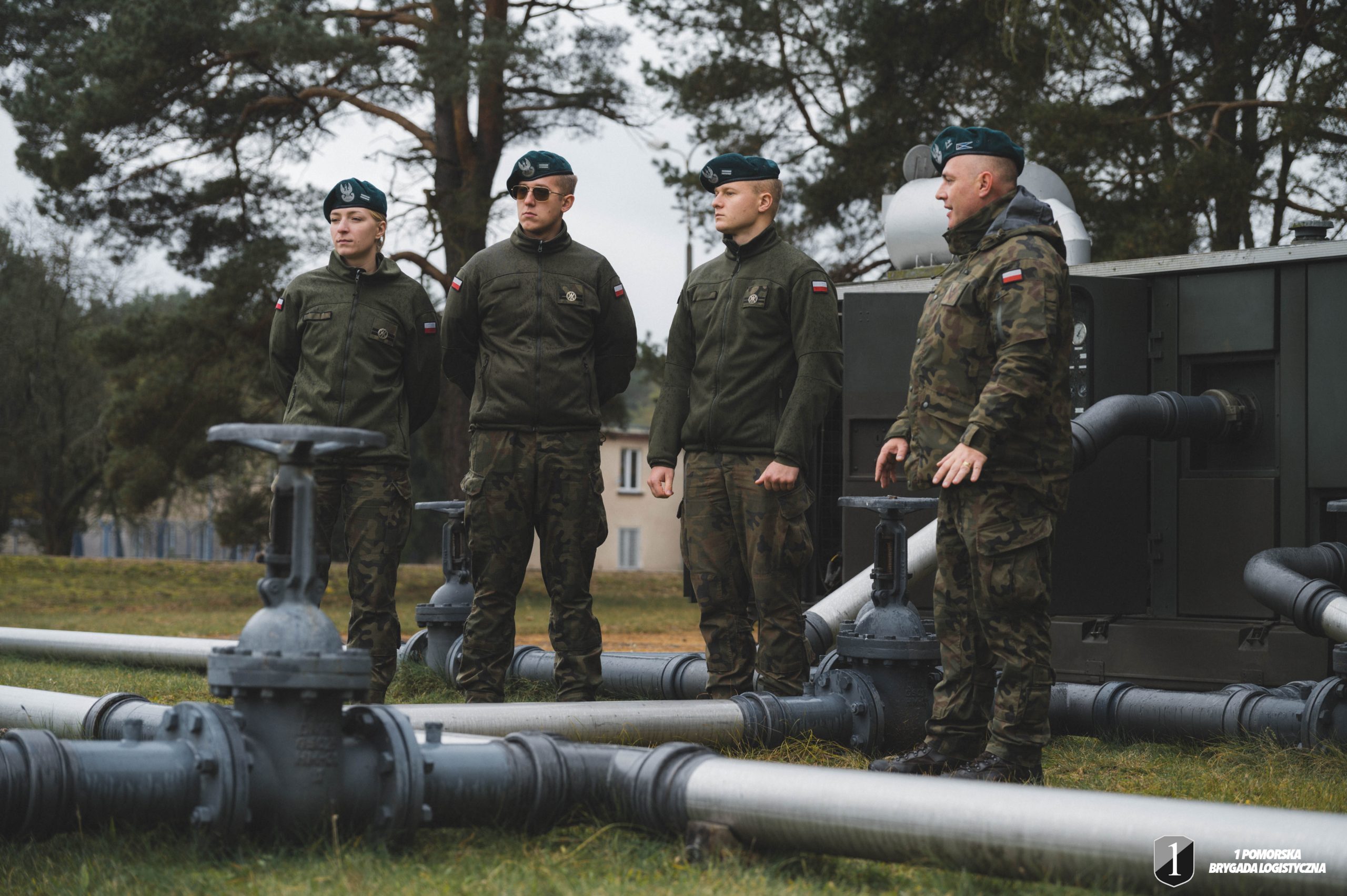 Podchorążowie przy początkowym punkcie rurociągu. Grupa żołnierzy stoi na dworzu przy rurociągi.