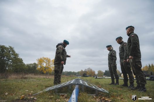 Podchorąży i prowadzący zajęcia stoją przy pomoście do pokonywania drogi utwardzonej nad rurociągiem.