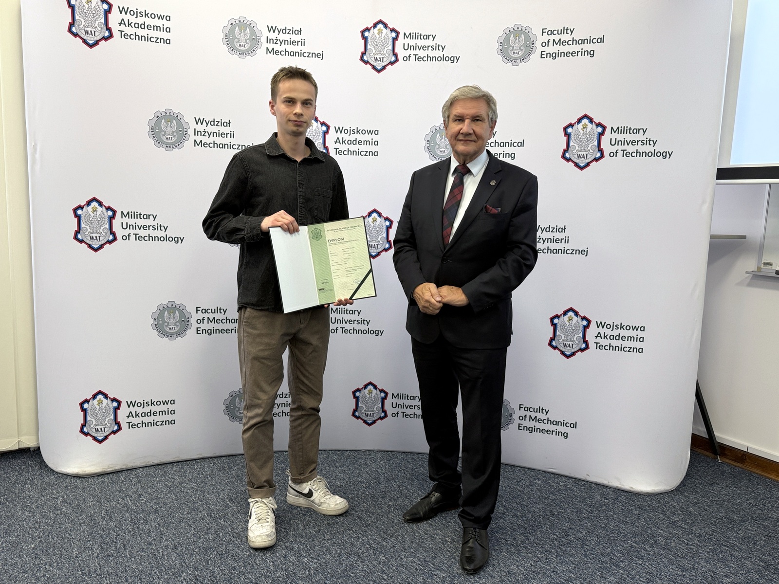 Dziekan wraz ze studentem trzymającym dyplom, w tle ścianka z logami WAT i WIM