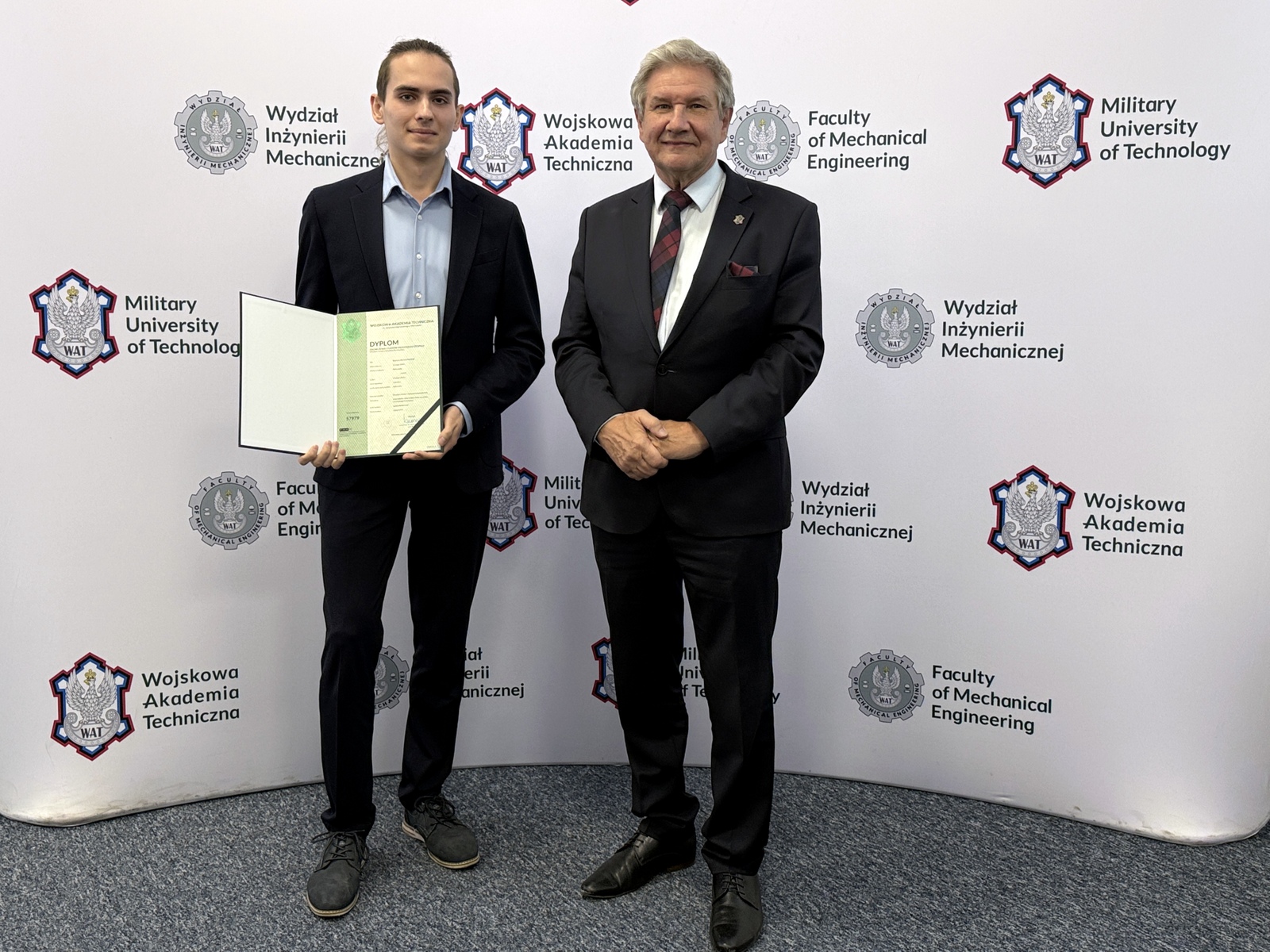 Dziekan wraz ze studentem trzymającym dyplom, w tle ścianka z logami WAT i WIM