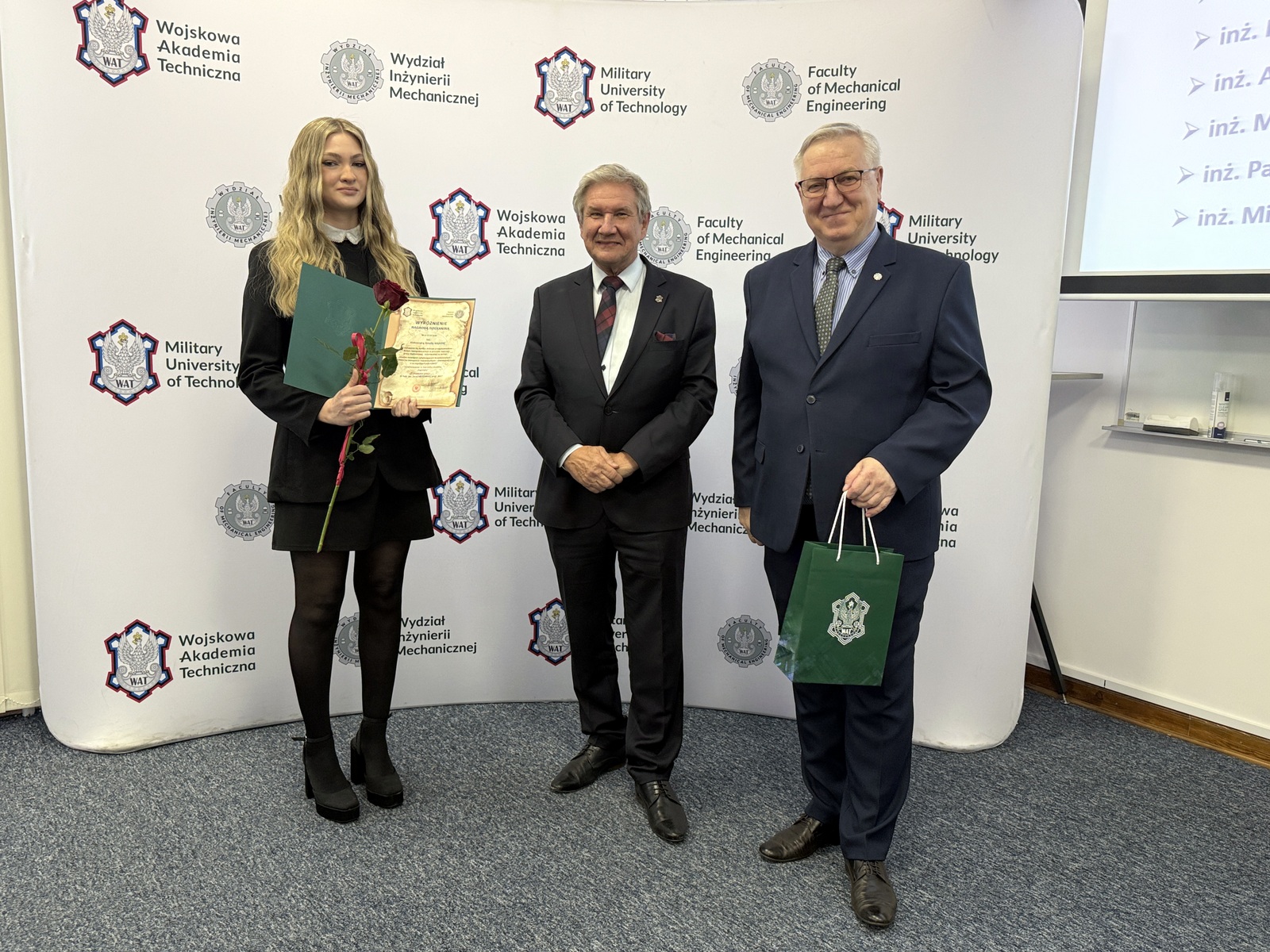 Dziekan oraz Kierownik Dziekanatu wraz ze studentką trzymającą wyróżnienie, w tle ścianka z logami WAT i WIM