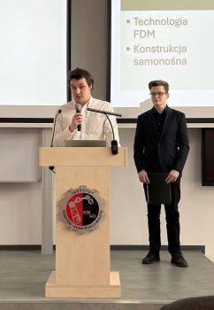 Dwóch studentów ubranych odświętnie podczas prezentacji pracy. Jeden trzyma mikrofon i stoi przy mównicy, drugi stoi za nim z teczką w rękach.