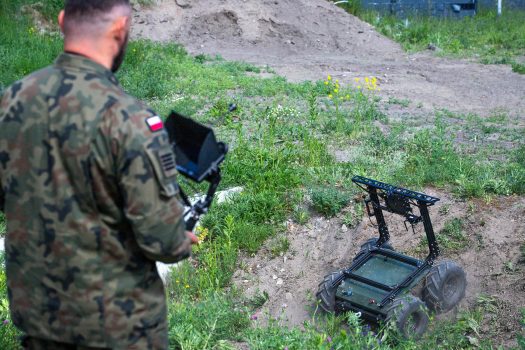 Widok od tyłu: operator wojskowy w mundurze steruje pulpitem; przed nim robot kołowy wyjeżdża z zagłębienia terenu.