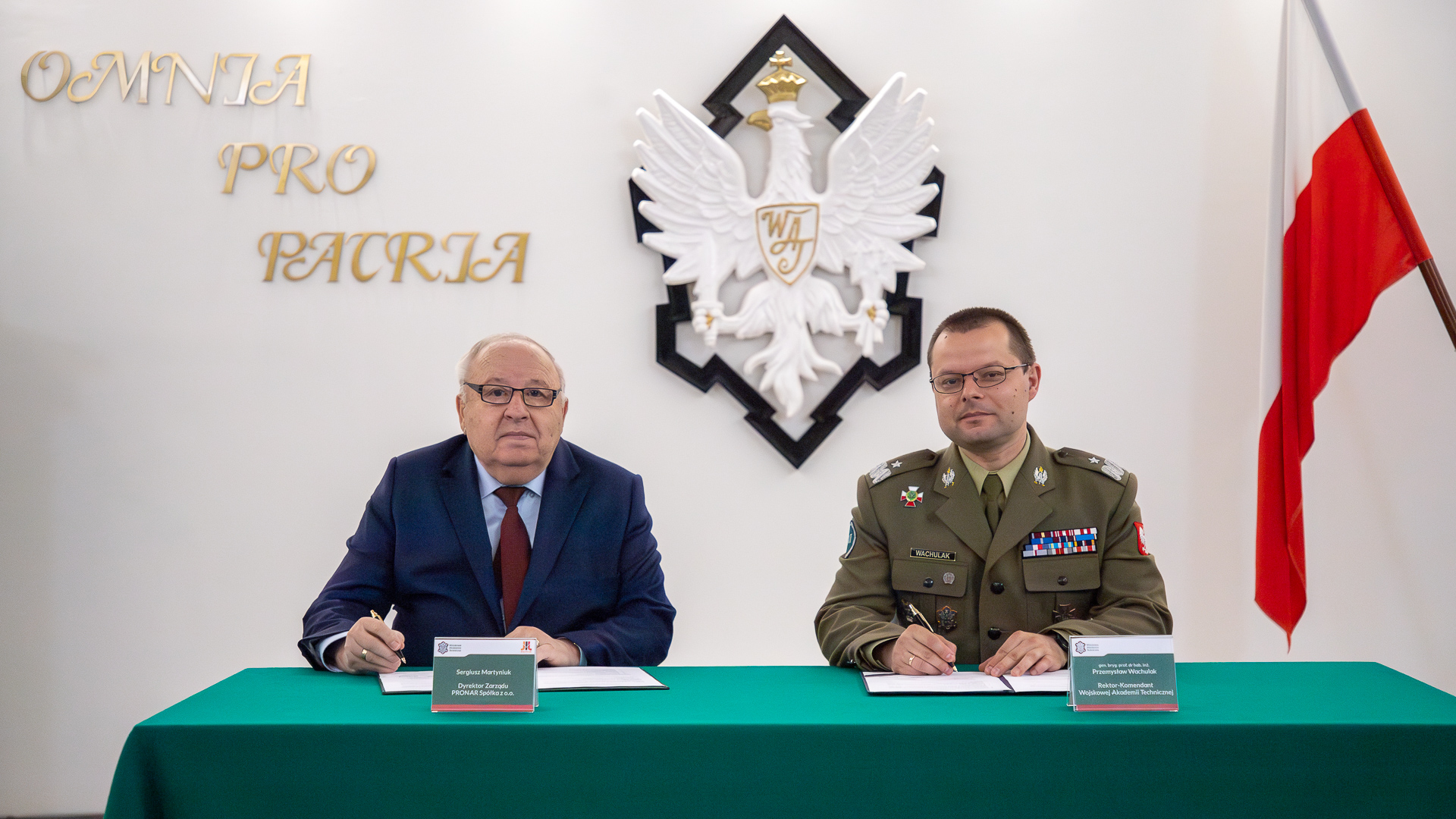 Rektor WAT i Dyrektor Pronar siedzący przy stole, w ręku trzymają długopisy, na stole leżą dokumenty, które Panowie pospisują. W tle flaga Polski i godło WAT.