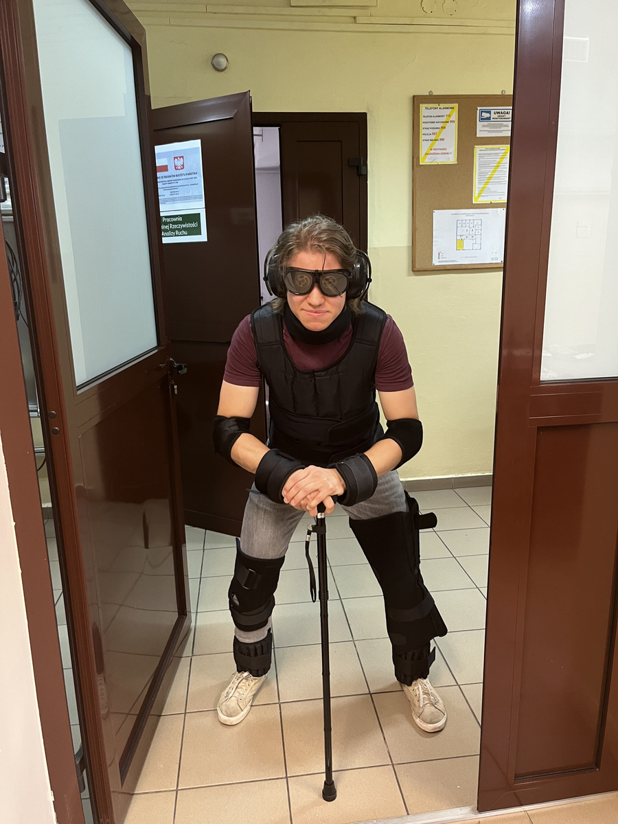Młoda kobieta, która ma założone elementy kostiumu do symulacji starości, także w specjalnych okularach, ręką podpiera laskę. Stoi w przejśiu między pomieszczeniami.