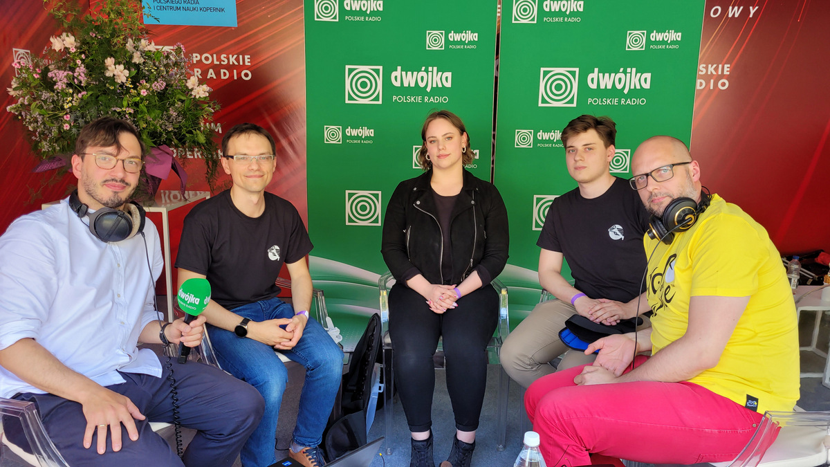 Przedstawiciele Koła Naukowego oraz redaktorzy radiowej Dwójki siedzą na krzesłach, w tle banery z nazwą stacji radiowej.