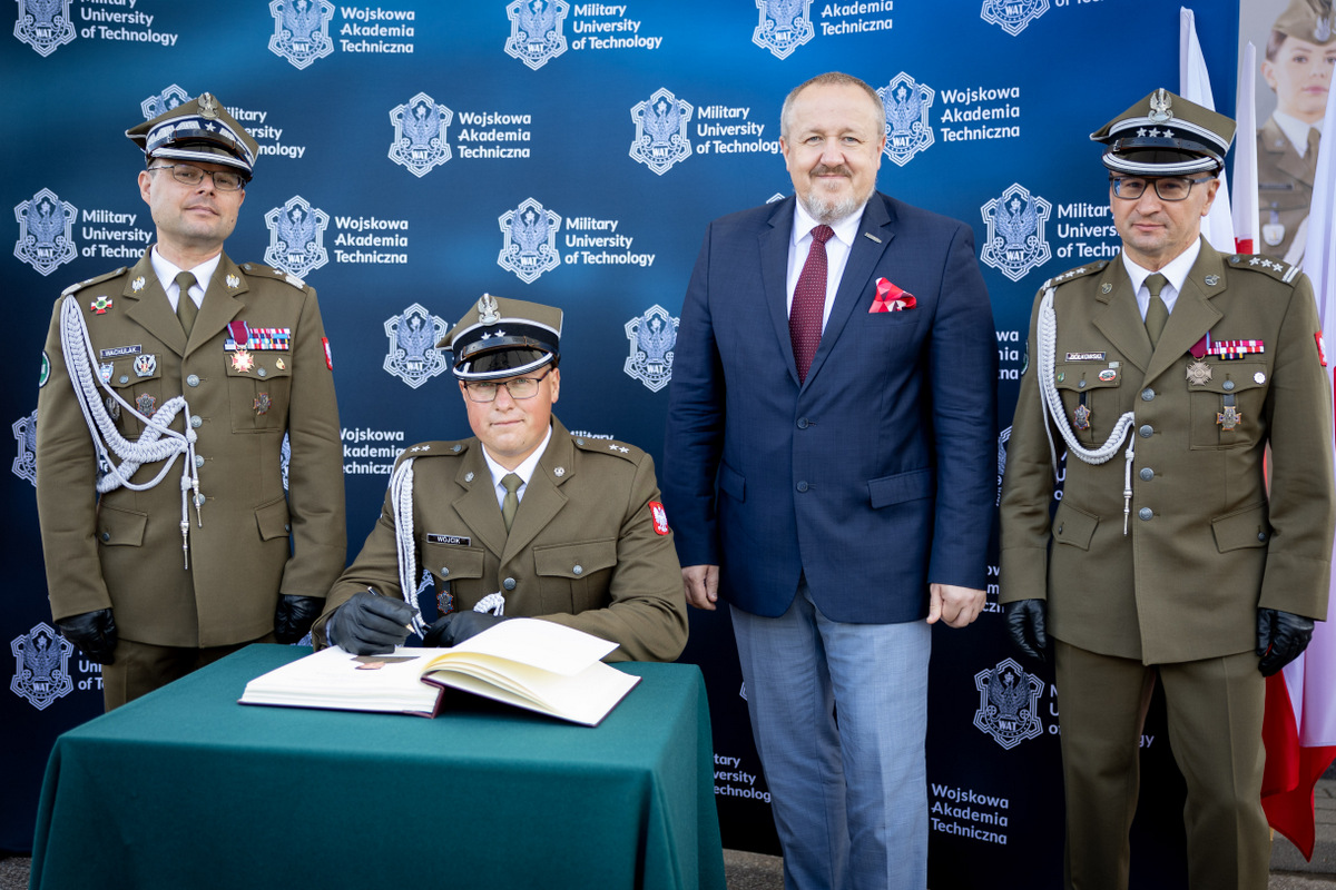 Na zdjęciu: Rektor-Komendant WAT, ppor. mgr inż. Adam Wójcik, Dziekan WIM, Zastępca Dziekana WIM. Młody oficer siedzi przy małym stoliku robiąc wpis w Księdze Pamiątkowej uczelni. Obok stoją pozostałe osoby.