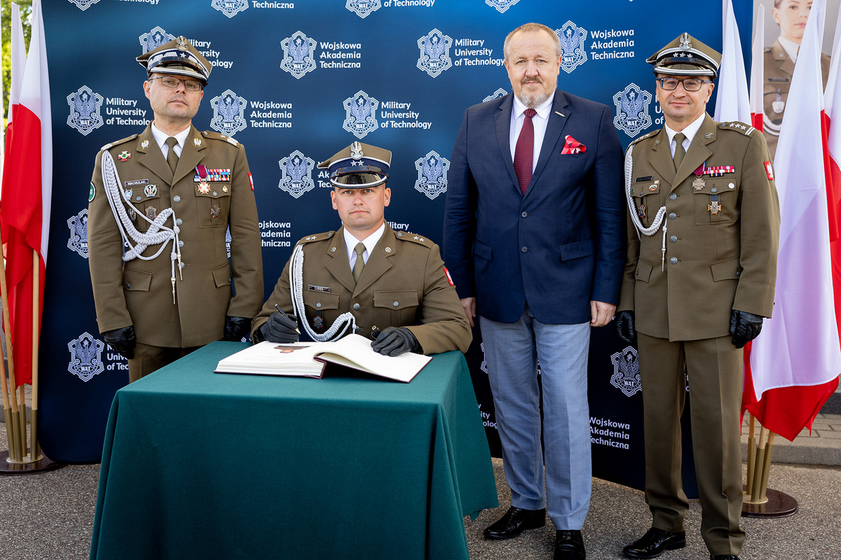 Na zdjęciu: Rektor-Komendant WAT, ppor. mgr inż. Adam Ciseł, Dziekan WIM, Zastępca Dziekana WIM. Młody oficer siedzi przy małym stoliku robiąc wpis w Księdze Pamiątkowej uczelni. Obok stoją pozostałe osoby.
