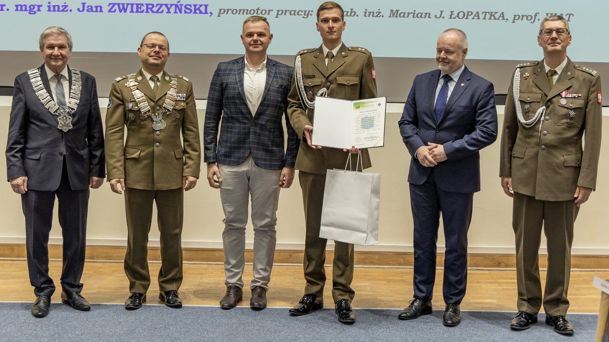 Tyłem do sceny, przodem do zgromadzonych stoi wyróżniona osoba wraz z osobami wręcającymi dyplom i upominki.