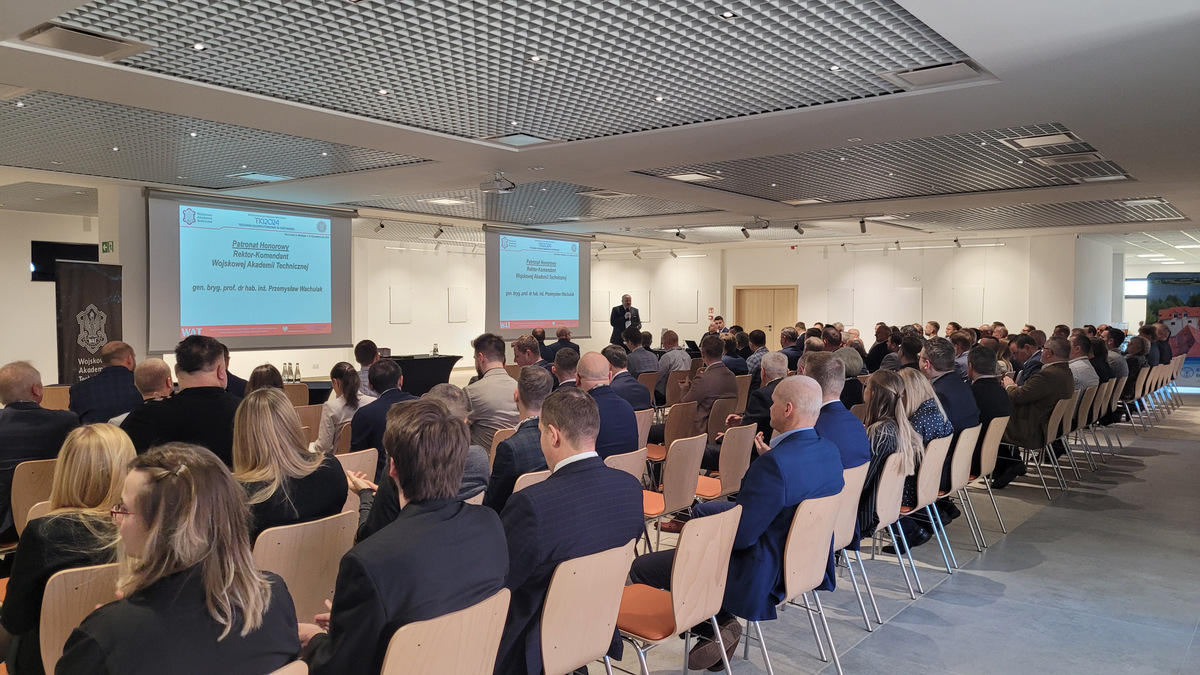 Powitanie gości przez prof. J. Małąchowskiego, stoim przodem do zgromadzonych na konferencji osób. Uczestnicy siedzą na krzesłach przodem do ekranów.